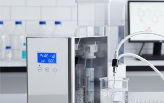 Laboratuvar ortamında çalışan dijital ekranlı saf su üretim cihazı ve cam behere doldurulan saf suyun detaylı yakın çekim görüntüsü.